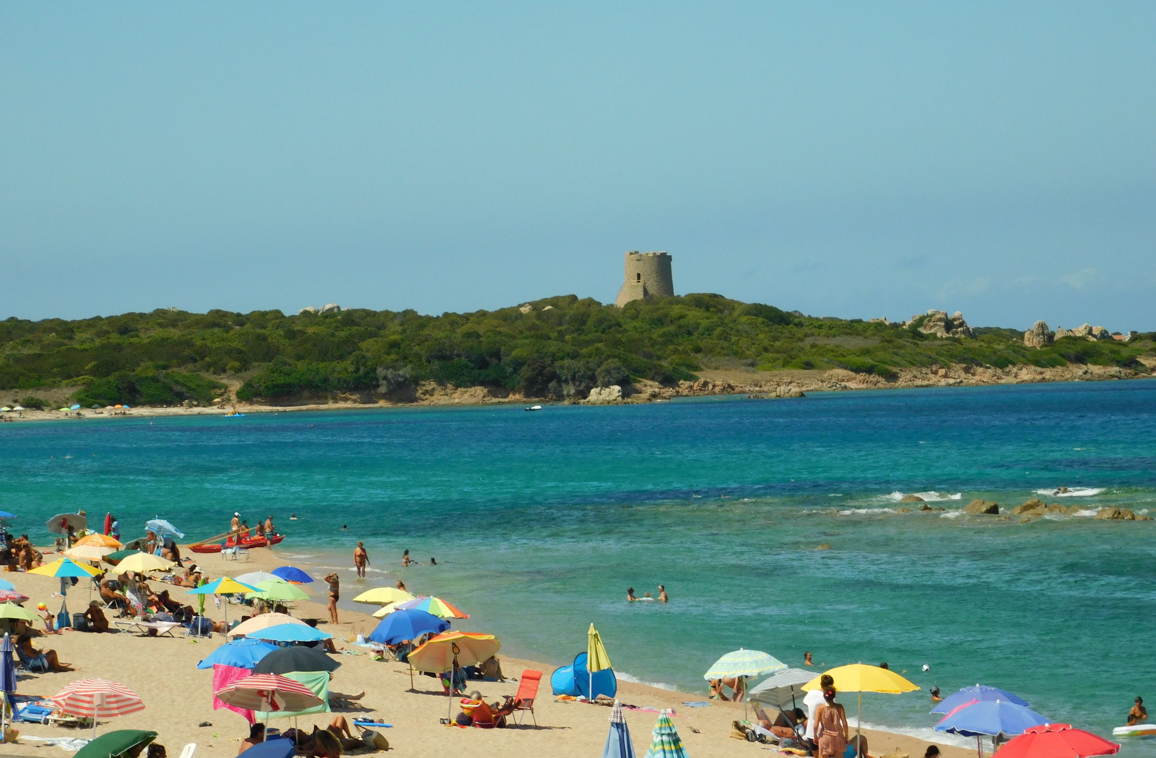 Une plage où des gens se prélassent au soleil et se baignent, avec des parasols, et un château perché sur une colline en arrière-plan.