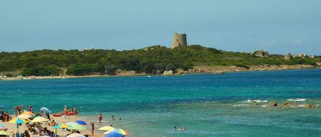A beach with people sunbathing and swimming, umbrellas, and a castle on a hill in the background.
