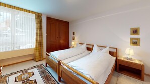 Habitación de hotel con dos camas, cabeceros de madera, un ventanal con cortinas translúcidas, un armario de madera y una alfombra estampada.