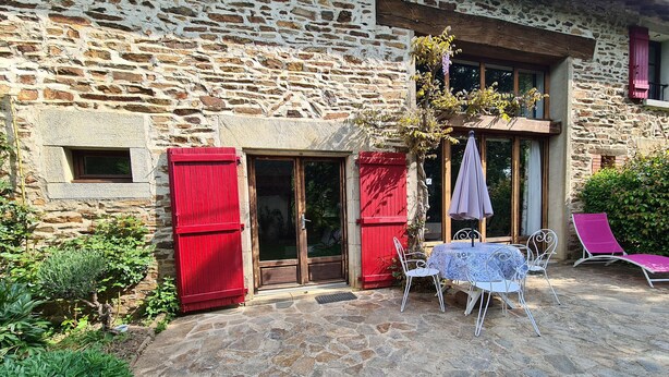 Ein Steinhaus mit roten Fensterläden, einer Glastür und einer Terrasse mit Tisch und Stühlen.