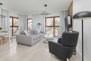 Un salon moderne avec un canapé gris, un fauteuil gris, une table à manger avec des chaises, un téléviseur et de grandes fenêtres.