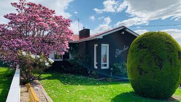 Una casa con fachada oscura, chimenea visible y un jardín bien cuidado con un arbusto grande y un árbol floreciente.