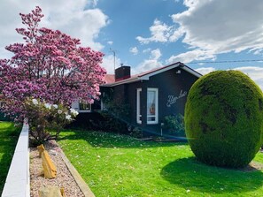 A house with a dark facade, a visible chimney, and a well-maintained garden with a large bush and flowering tree.