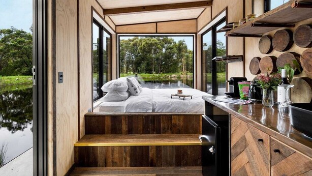 A modern, compact bedroom with a wooden floor, a bed with white linens, and a view of a lake through large windows. - Getawayz NSW - 'Banksia' Designer Tiny Home w/ stunning dam views (Kangaroo Valley)
