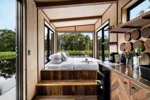 Een moderne, compacte slaapkamer met een houten vloer, een bed met witte lakens en uitzicht op een meer door grote ramen.