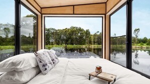 A bedroom with a large bed, a wooden bedside table, and a view of a lake and trees. - Getawayz NSW - 'Banksia' Designer Tiny Home w/ stunning dam views (Kangaroo Valley)