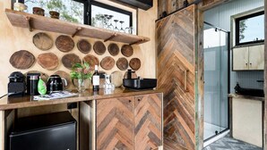 A compact kitchen with a wooden herringbone floor, a black refrigerator, and a wooden shelf with jars and glasses. - Getawayz NSW - 'Banksia' Designer Tiny Home w/ stunning dam views (Kangaroo Valley)