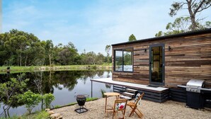 A modern cabin with a wooden exterior, a deck, a grill, and a view of a lake. - Getawayz NSW - 'Banksia' Designer Tiny Home w/ stunning dam views (Kangaroo Valley)