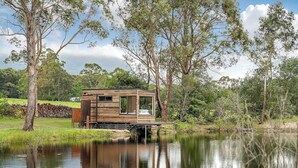 A wooden cabin with a deck, situated by a calm lake surrounded by trees. - Getawayz NSW - 'Banksia' Designer Tiny Home w/ stunning dam views (Kangaroo Valley)