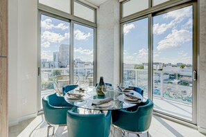 A modern dining area with a glass table and blue chairs, overlooking a cityscape through large windows.