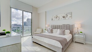 Un dormitorio moderno con una cama grande, vistas a la ciudad, un escritorio blanco y una mesita de noche con lámparas.