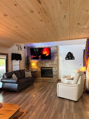 Ein Wohnzimmer mit Kamin aus Stein, einem Flachbildfernseher, einem Ledersofa, einem Liegestuhl und einer Holzbalkendecke.