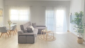 A modern living room with a grey sectional sofa, a small round table, and a dining area with a glass table and chairs. - HomeForGuest Two bedroom Apartment with pool and terrace (Antigua - Fuerteventura)