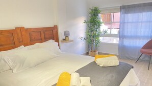 A neatly made bed with white and yellow towels, a wooden headboard, a bedside plant, and a window with a view of buildings. - HomeForGuest Two bedroom Apartment with pool and terrace (Antigua - Fuerteventura)