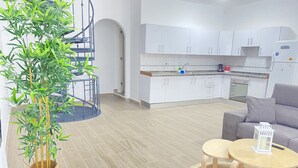 A modern kitchen with white cabinets, a built-in oven, and a coffee machine. There is a grey sofa and a small round table with a lantern. A large potted plant is in the corner. - HomeForGuest Two bedroom Apartment with pool and terrace (Antigua - Fuerteventura)