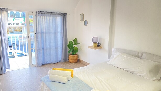 A hotel room with a bed, a potted plant, a bedside table with a lamp, and a view of the outside through a door. - HomeForGuest Two bedroom Apartment with pool and terrace (Antigua - Fuerteventura)
