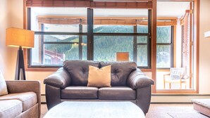 Ein Zimmer mit Ledersofa, Holztisch und Blick auf Berge durch große Fenster.