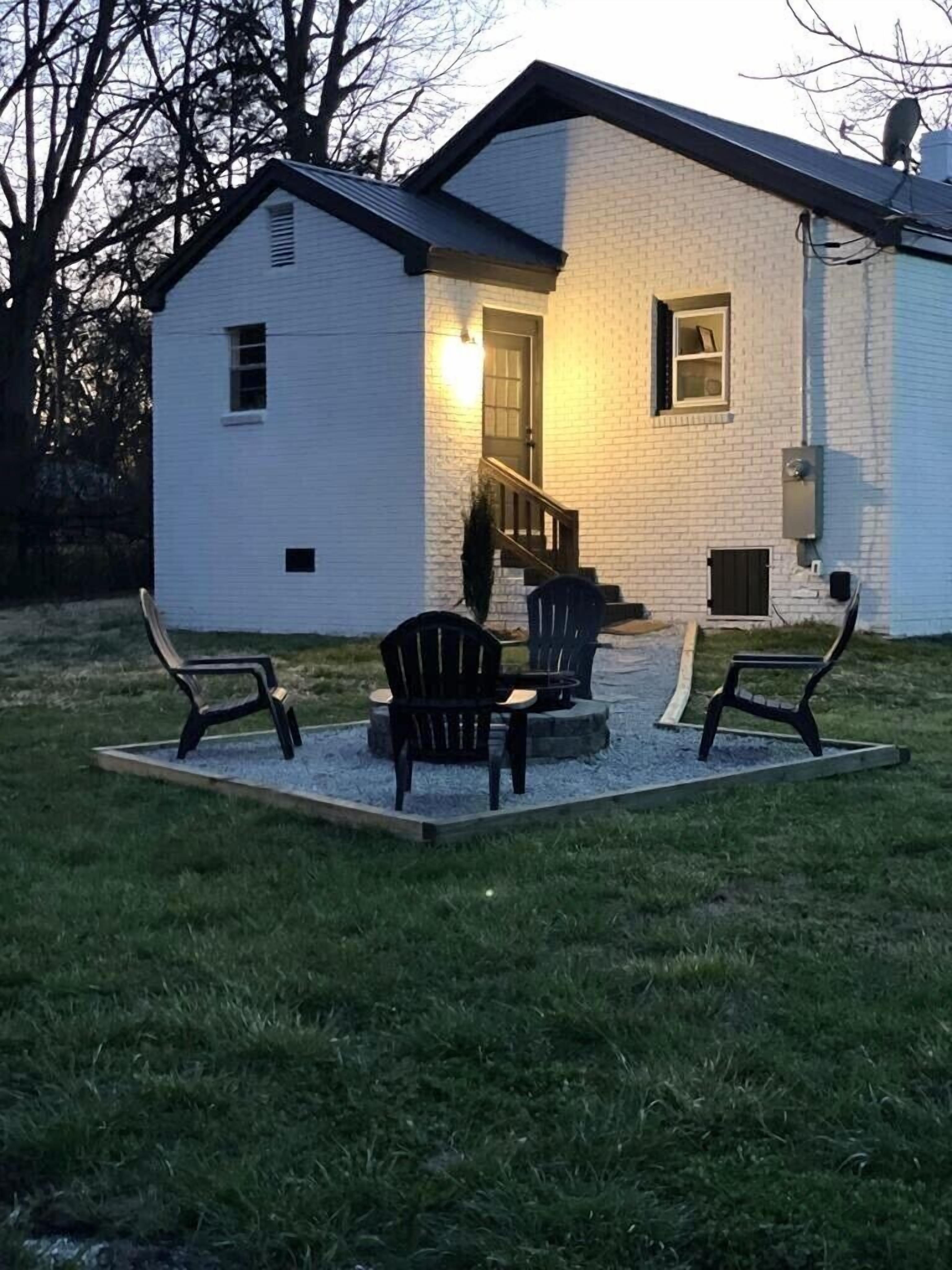 Una casa de ladrillo blanco con un porche cubierto, dos sillas Adirondack y una pequeña mesa.