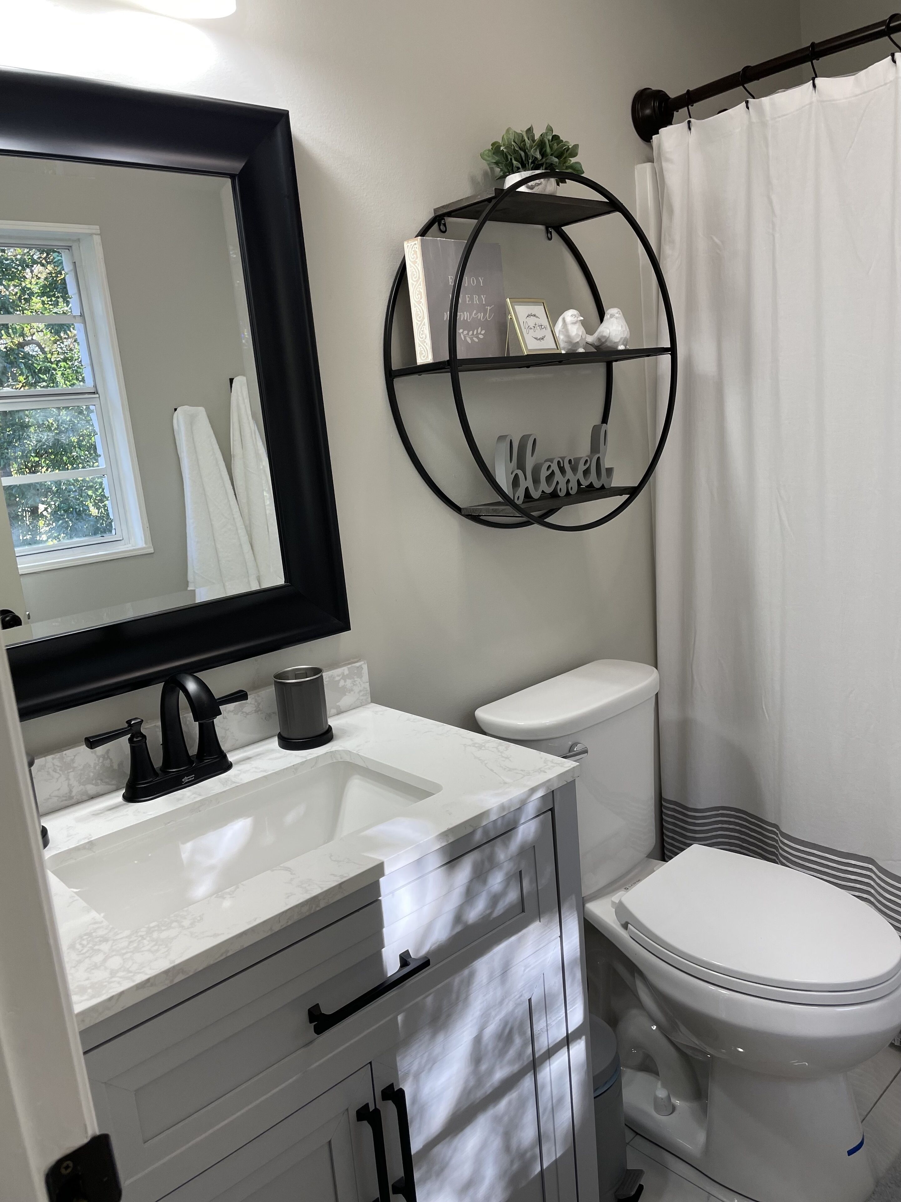 A bathroom with a white sink, black faucet, and a toilet. There is a circular shelf with decorative items and a sign that says "Blessed".