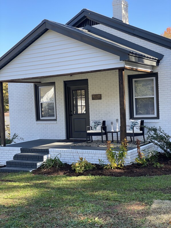 A single-story house with a white exterior, a black door, and a 'WELCOME' sign. There are two chairs and a small table on the porch. - Private, pet-friendly adorable getaway mins to downtown (Greenville)