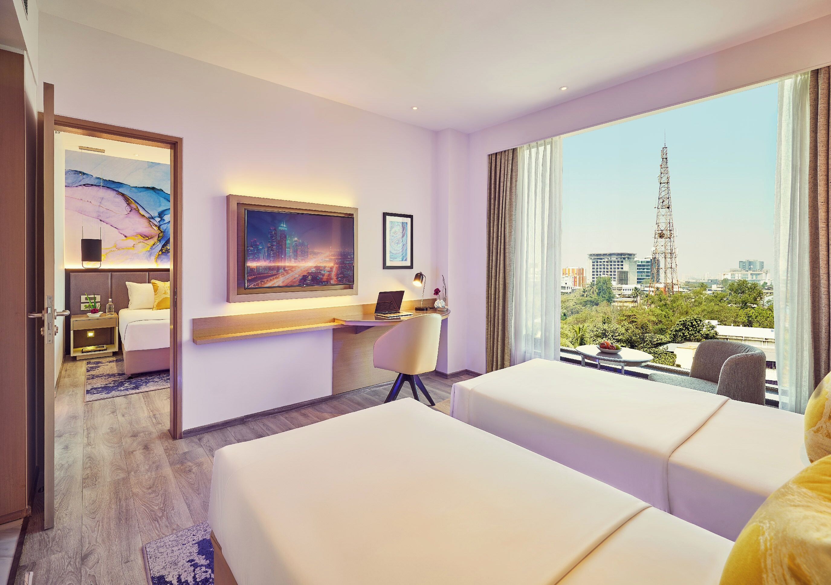 A hotel room with two beds, a desk with a laptop, a large window with a city view, and artwork on the walls.