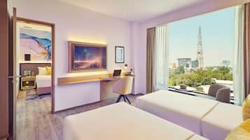 A hotel room with two beds, a desk with a laptop, a large window with a city view, and artwork on the walls.