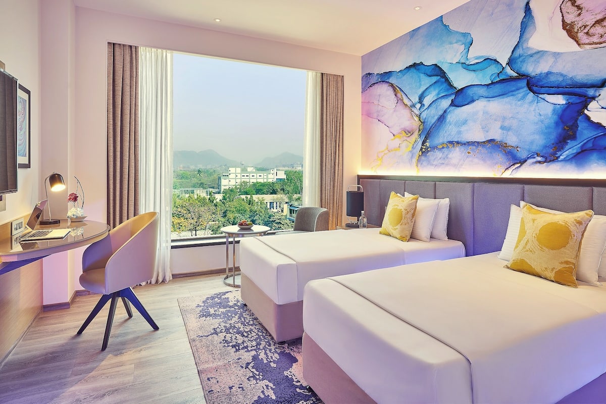 Habitació d'hotel amb escriptori, dos llits, un quadre abstracte gran i vista a la ciutat.