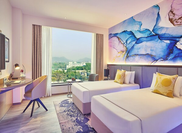 Une chambre d’hôtel avec un bureau, deux lits, un grand tableau abstrait et une vue sur la ville.
