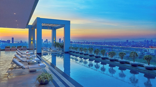 Un área de piscina en la azotea con sillones de descanso y plantas en macetas, con vistas al horizonte de la ciudad al atardecer.