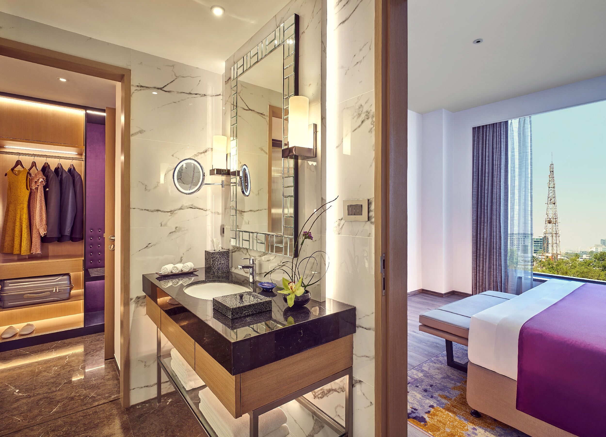A hotel room with a marble bathroom, a vanity with a mirror, and a bed with a view of the city.