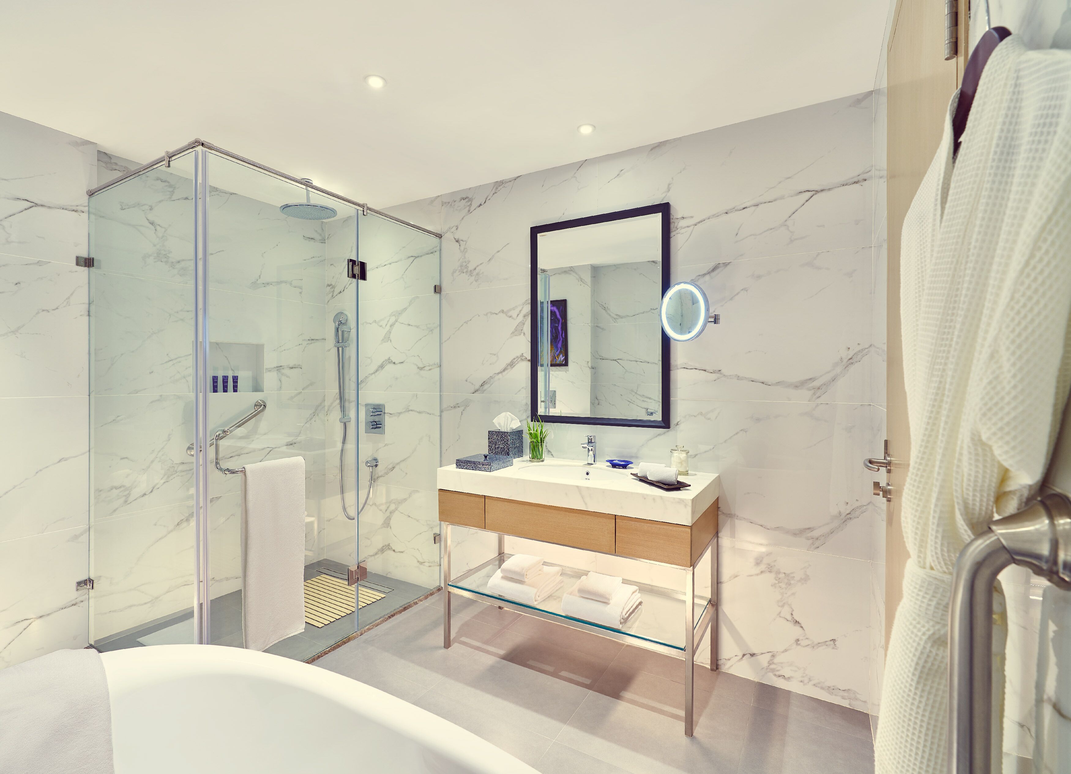 Un baño moderno con ducha de vidrio, pared de azulejos de mármol, tocador de madera y un espejo grande con luz redonda.