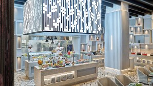 Un restaurante moderno con una barra central que exhibe diversas opciones de comida, rodeado de zonas de estar con mesas y sillas.