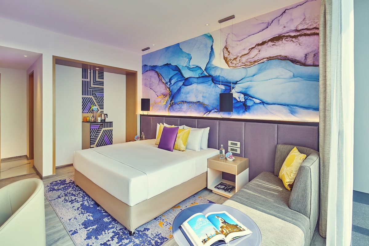 Una habitació d'hotel moderna amb un llit gran, una zona de seients, una paret amb art abstracte i una taula petita amb un llibre.