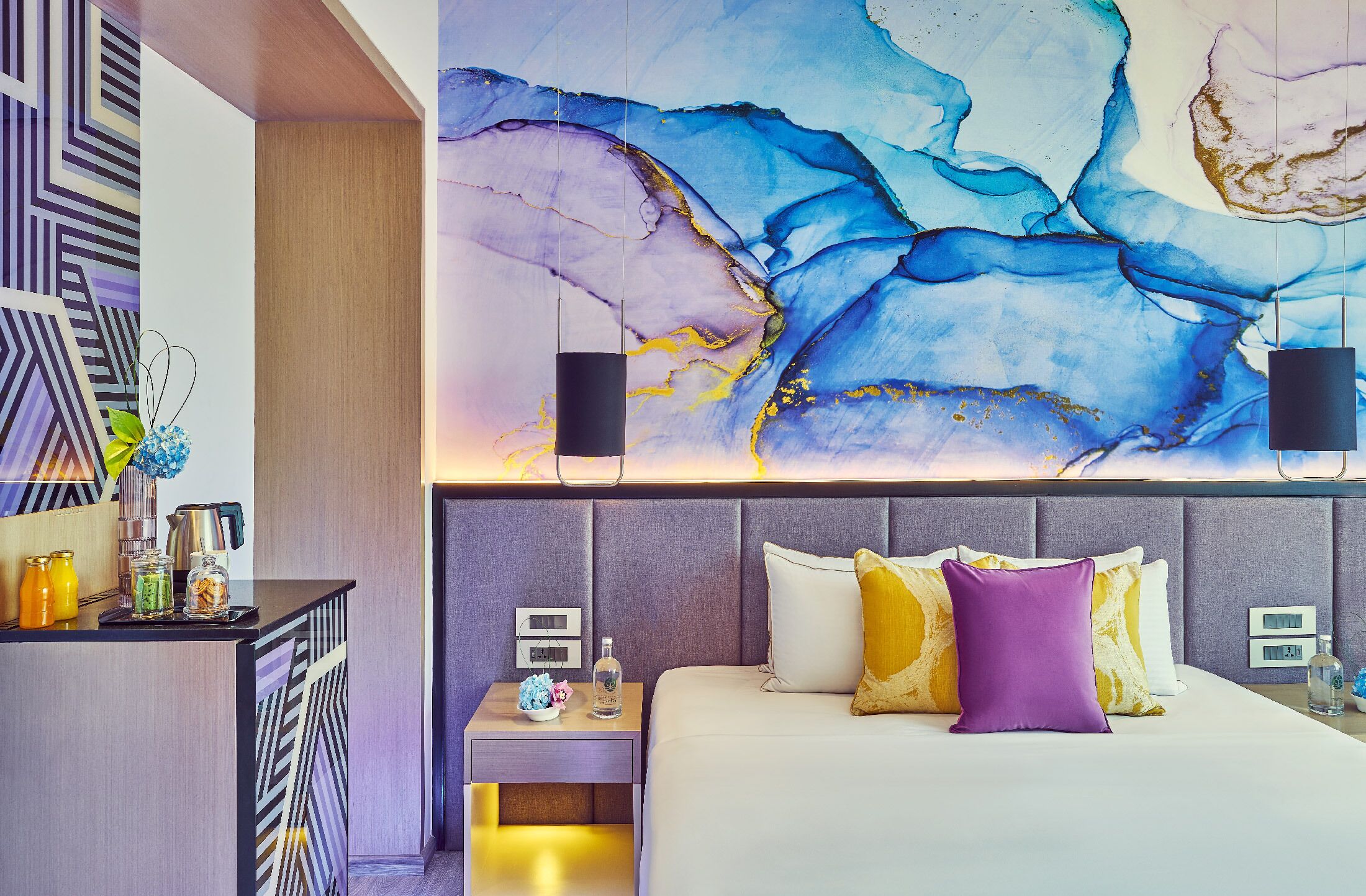 Una habitación de hotel moderna con un cuadro abstracto grande en la pared, una cama bien hecha con muchos cojines y una mesita de noche pequeña con una lámpara y una botella.