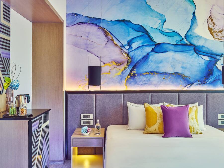 Una habitación de hotel moderna con un cuadro abstracto grande en la pared, una cama bien hecha con muchos cojines y una mesita de noche pequeña con una lámpara y una botella.