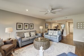 A living room with a grey sofa, a coffee table, and a patterned rug. There is a ceiling fan and framed pictures on the wall.
