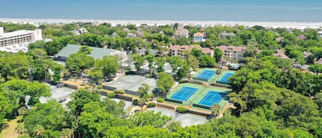 Ett flygbildsbild av ett bostadsområde med flera tennisbanor omgivna av frodig grönska, en strand i bakgrunden och en blandning av en- och flerfamiljshus.
