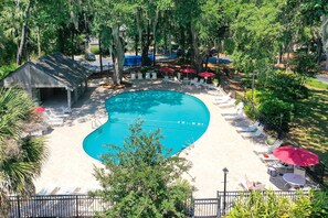 Una piscina con sillas de descanso y sombrillas, rodeada de árboles y una zona cubierta.