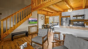 A spacious living area with a wooden staircase, a dining table with chairs, a kitchen with modern appliances, and a large TV on the wall. - Rave ReVIEWs! HOA Pool! Fire Pit! Hot Tub! Arcade! (Gatlinburg)