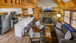 A spacious kitchen with modern appliances, a dining area with wooden chairs, a stone fireplace, and a cozy seating area with a sofa and cushions. - Rave ReVIEWs! HOA Pool! Fire Pit! Hot Tub! Arcade! (Gatlinburg)