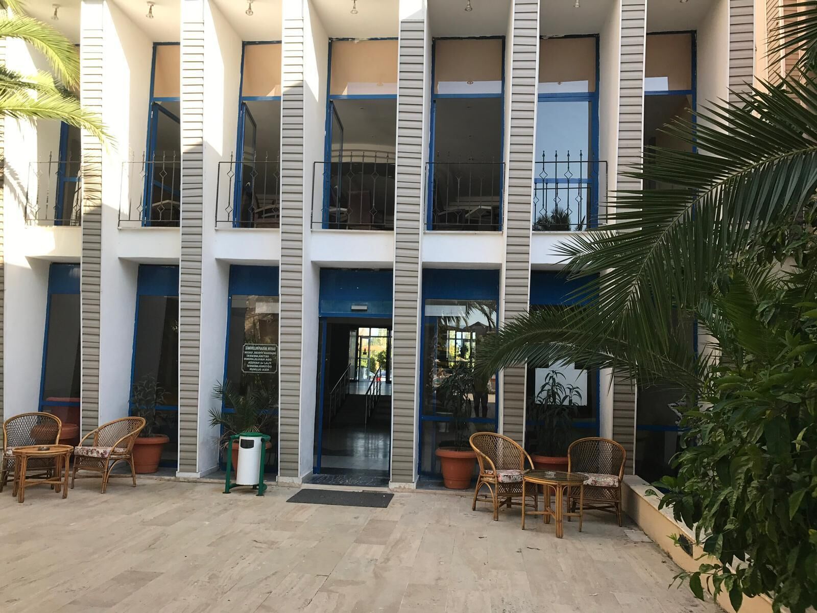 A multi-story building with a central entrance, outdoor seating area with wicker chairs, and potted plants.
