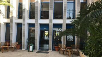 A multi-story building with a central entrance, outdoor seating area with wicker chairs, and potted plants.