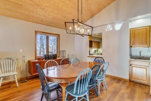 A dining room with a wooden table and chairs, a window with a view of trees, and a kitchen area with wooden cabinets. - Slopeside on Timberline Mountain's Salamander Trail-Perfect in every season! (Canaan Valley)