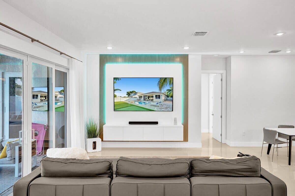 Een moderne woonkamer met een grote flatscreen televisie aan een witte muur, een grijze bank en een eethoek met een tafel en stoelen.