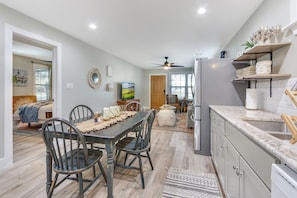A modern kitchen with a dining area, featuring a long table with chairs, a refrigerator, and a ceiling fan.