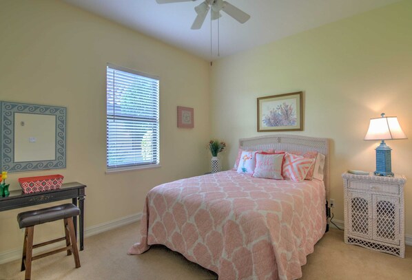 A bedroom with a bed, a desk, a chair, a window with blinds, a ceiling fan, a nightstand, a lamp, and a decorative wall piece.