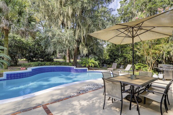 A swimming pool with blue tiles, surrounded by lush greenery and trees with hanging moss. There is a table with chairs and a large umbrella. - Pet and Friendly Shipyard Home with 5 Beds/3.5 Baths Firepit and Private Pool (Hilton Head)
