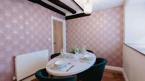 A dining area with a round table set for four, surrounded by dark chairs. A white radiator is visible on the left, and a door is seen in the background.