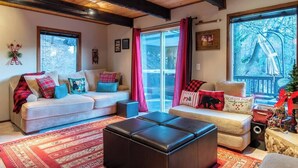 A cozy living room with a sofa, ottoman, and a large window with curtains. - Serene View Chalet | Hot Tub - King Beds Mtn Views (Big Bear Lake)
