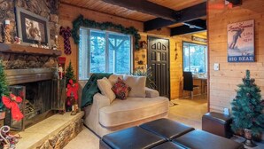 A cozy cabin interior with a fireplace, a couch, and a Christmas tree. - Serene View Chalet | Hot Tub - King Beds Mtn Views (Big Bear Lake)